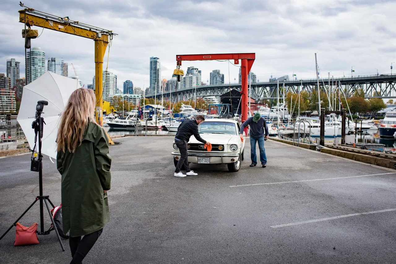 Photo crew preparing a white classic Ford Mustang beside a marina and bridge