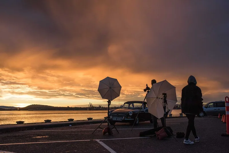 Photographers with studio umbrellas beside a classic car at a waterfront sunset